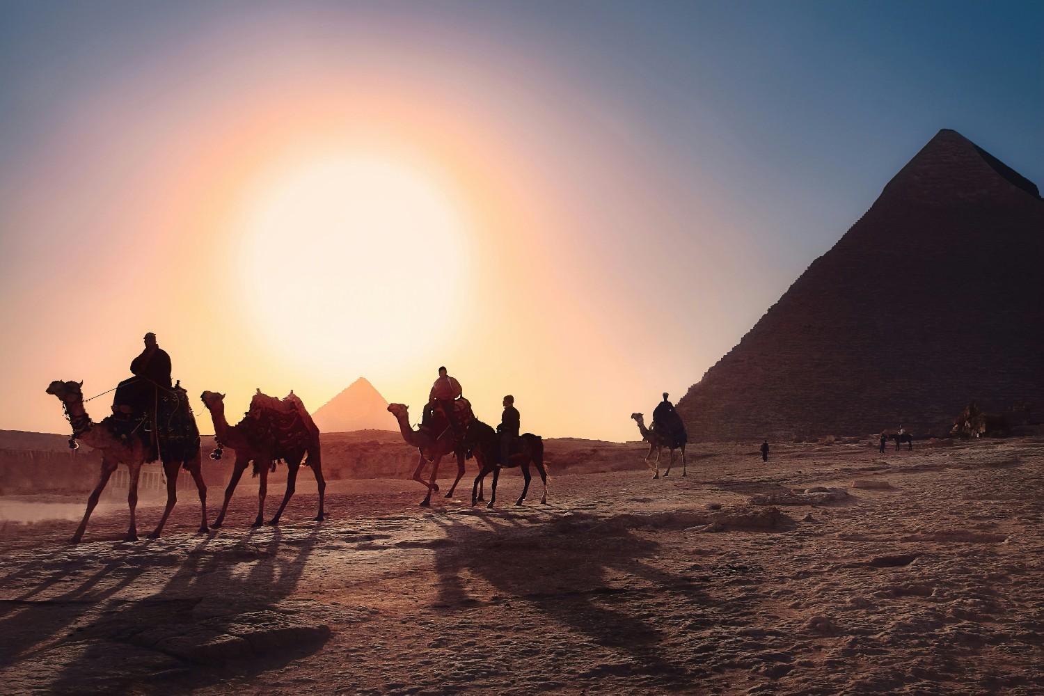 Tourists riding camels at sunset with pyramids silhouetted against golden sky, creating magical desert experience